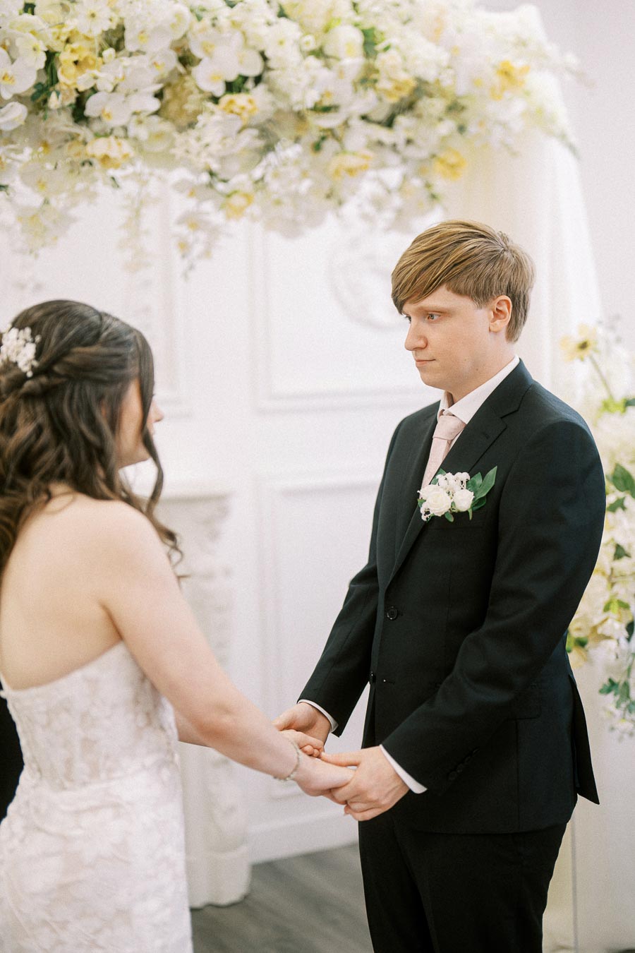 Bride and groom holding hands during a wedding ceremony in a beautifully decorated venue with white and yellow flowers.