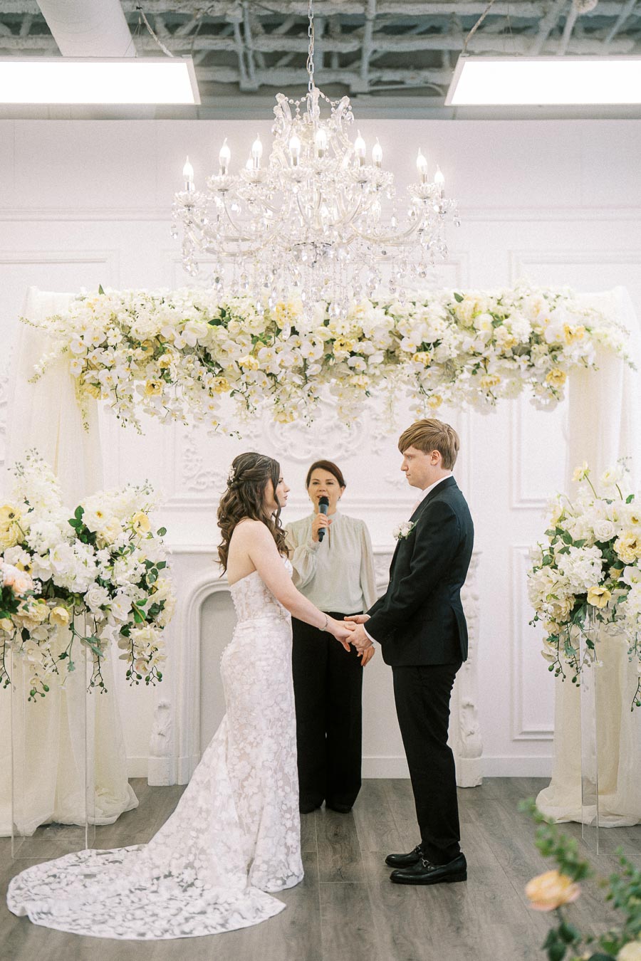 Elegant indoor wedding ceremony with bride and groom exchanging vows under a floral arch, with a chandelier overhead and an