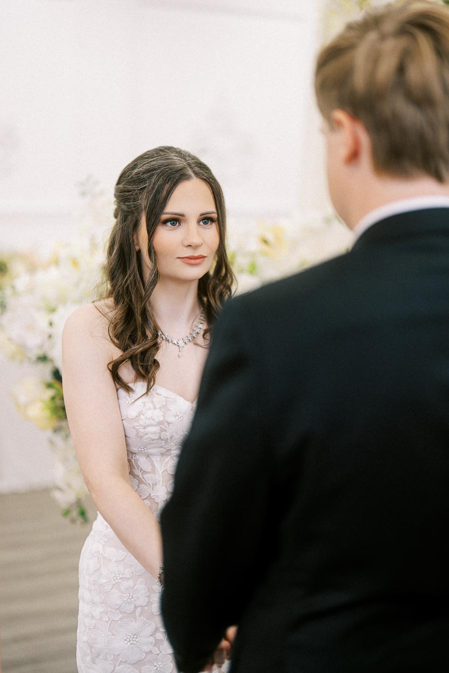 Bride and groom facing each other during a wedding ceremony, with the bride in a white floral lace dress and the groom in a