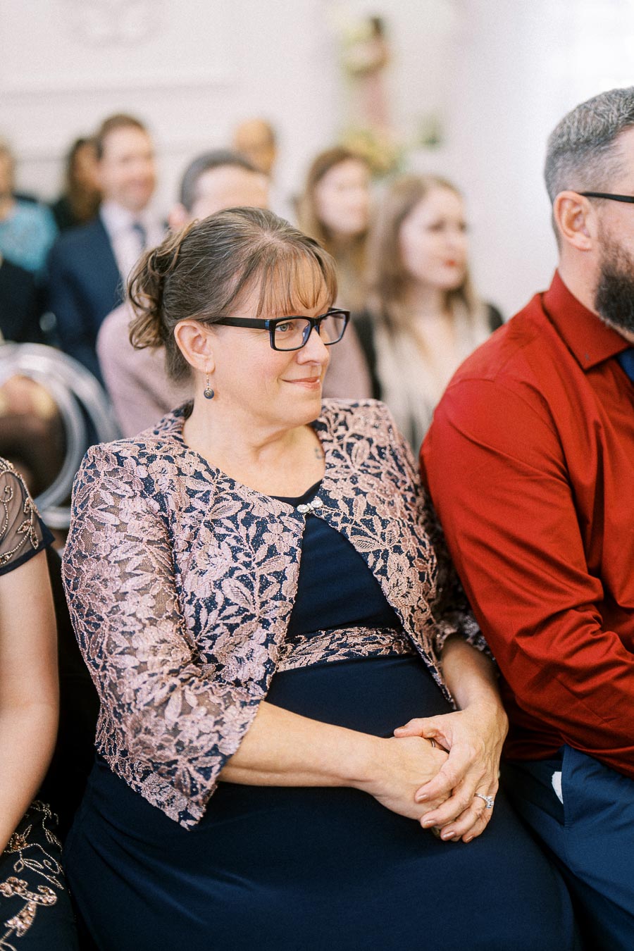 Middle-aged woman wearing glasses and a floral jacket at a formal event, seated next to a person in a red shirt, with other