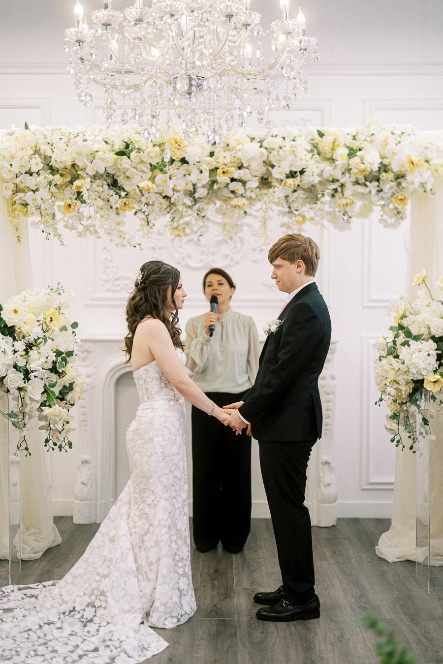 Bride and groom holding hands during wedding ceremony under a floral arch, with a crystal chandelier overhead and an