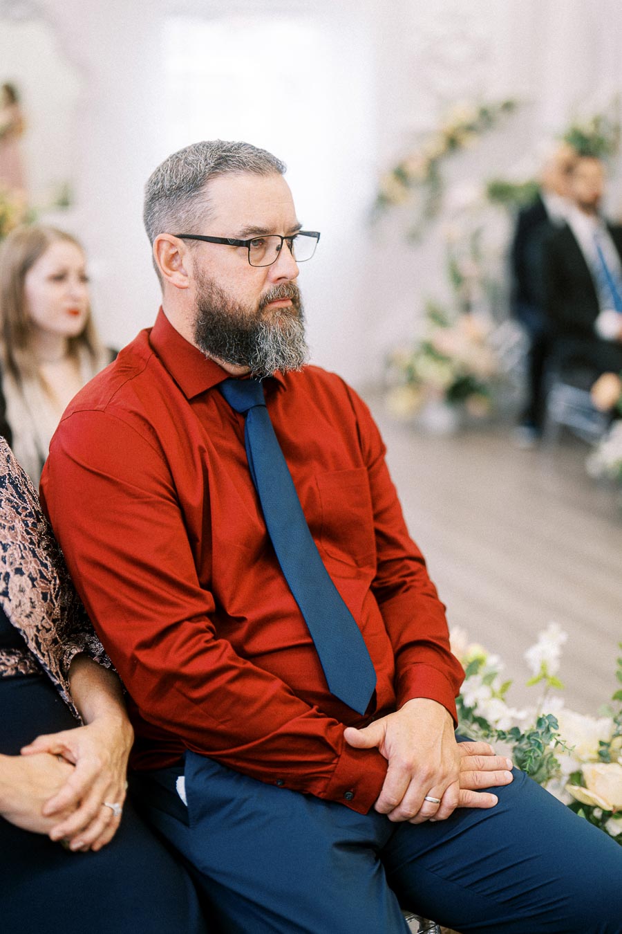 Man with a beard sitting attentively in an event, wearing a red shirt and blue tie, with blurred floral decorations in the
