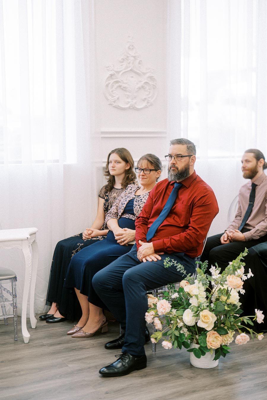 Family seated at a formal event, with a man in a red shirt and blue tie, and a floral arrangement nearby, inside a white,