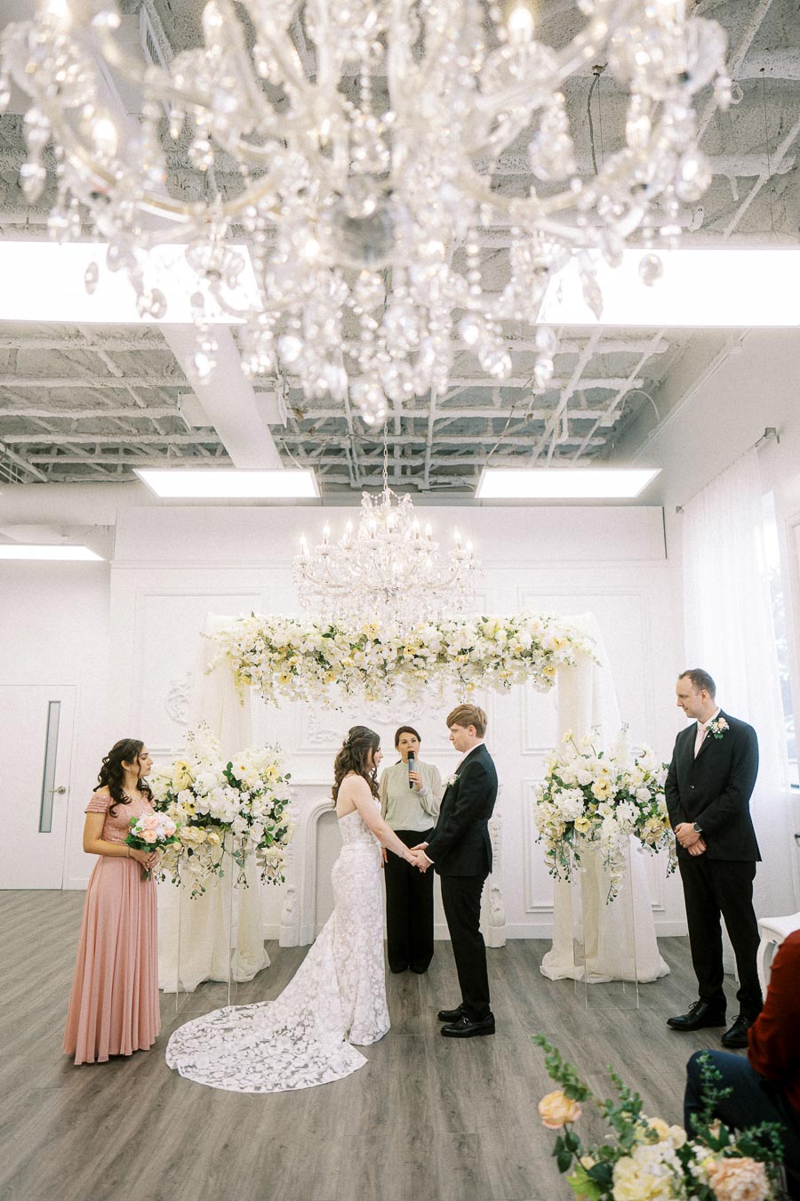 Indoor wedding ceremony with bride and groom holding hands under a floral arch, framed by elegant chandeliers and attended