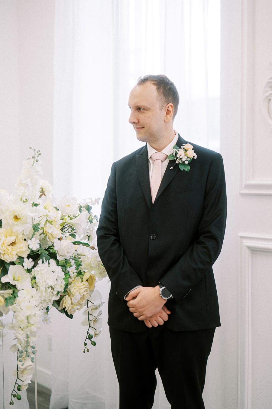 Man in a black suit with a pink tie and boutonniere stands inside a bright room with elegant flower arrangements, looking to