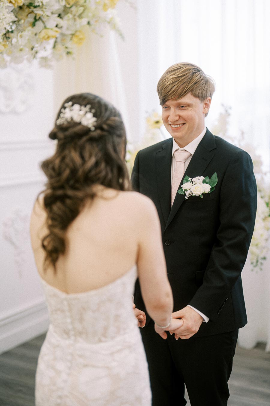 Wedding ceremony moment with bride and groom holding hands, bride in elegant lace gown and groom in black suit with floral