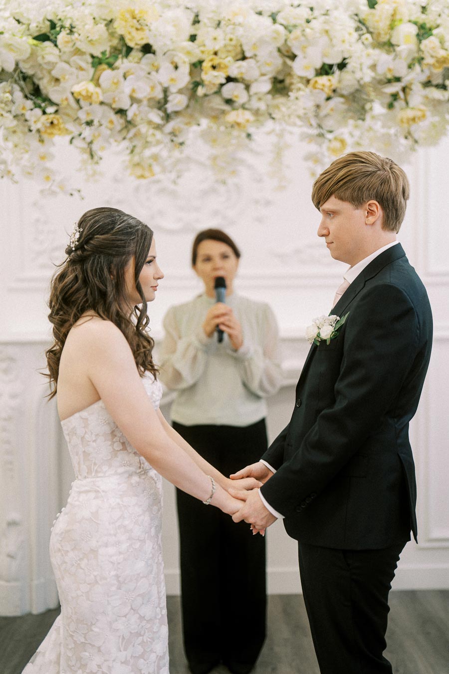 Bride and groom holding hands during their wedding ceremony, with a floral arrangement above and an officiant in the