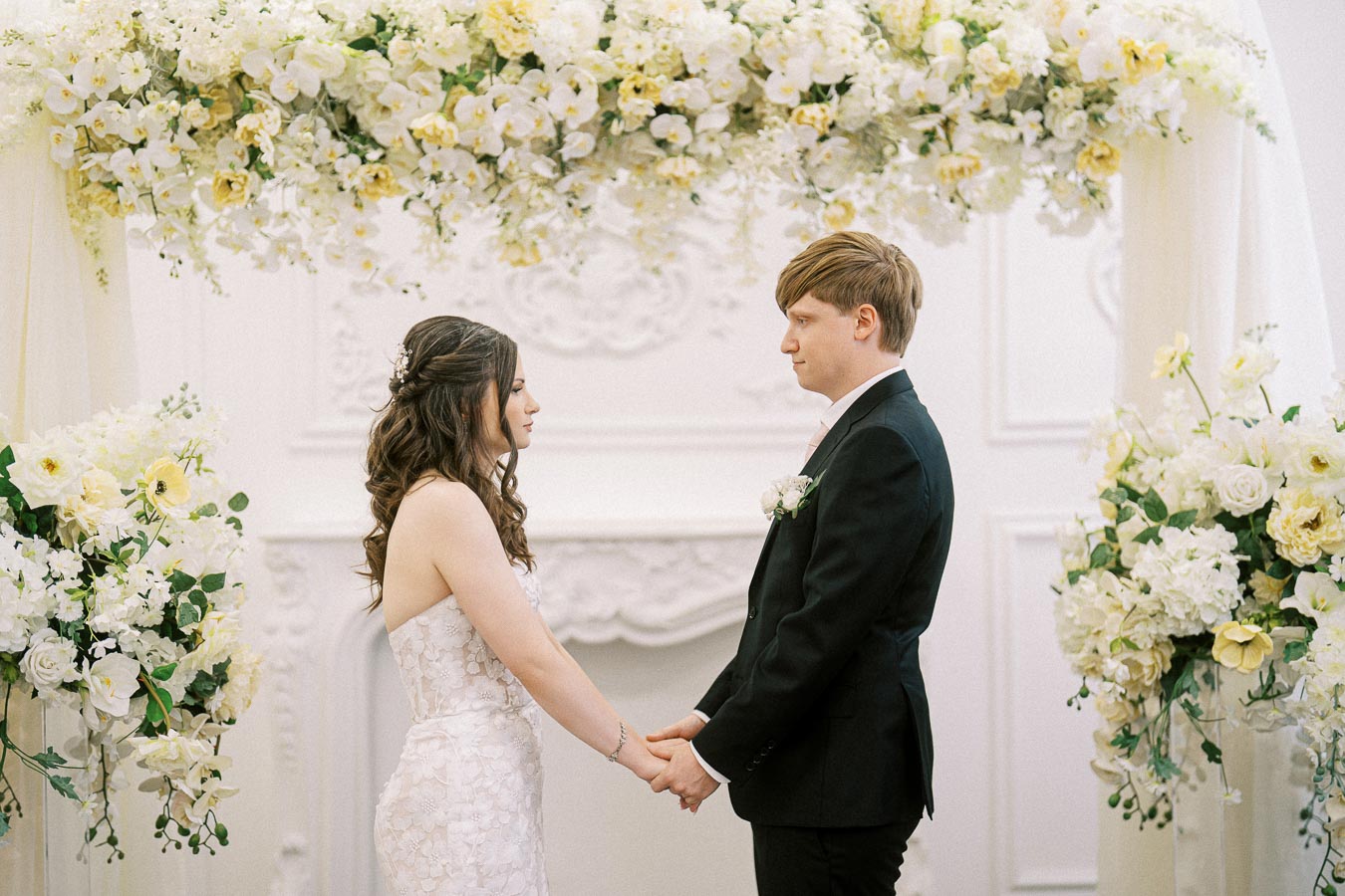 A bride and groom stand facing each other holding hands under an elegant floral arch adorned with white and yellow flowers,