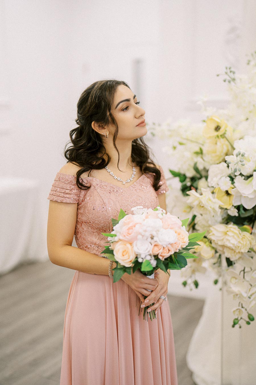 Bridesmaid in an elegant pink dress holding a bouquet of pastel flowers, standing near a floral arrangement.