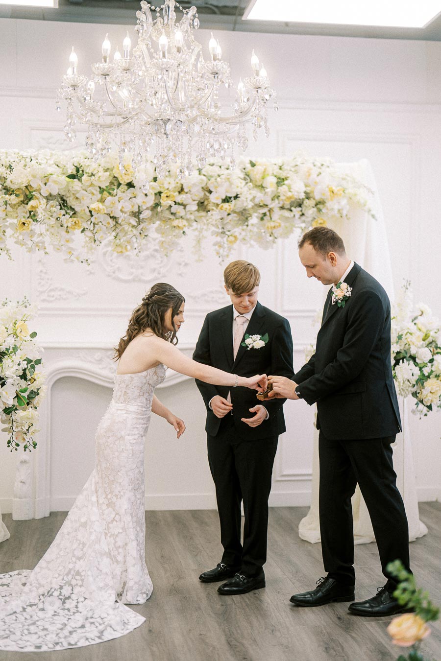 Elegant indoor wedding ceremony with a bride in a lace gown and two groomsmen in black suits exchanging rings under a floral