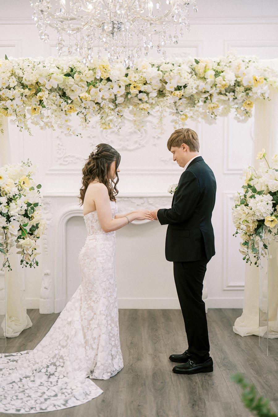 Elegant wedding ceremony with bride in a lace gown and groom in a black suit exchanging vows under a floral arch with white