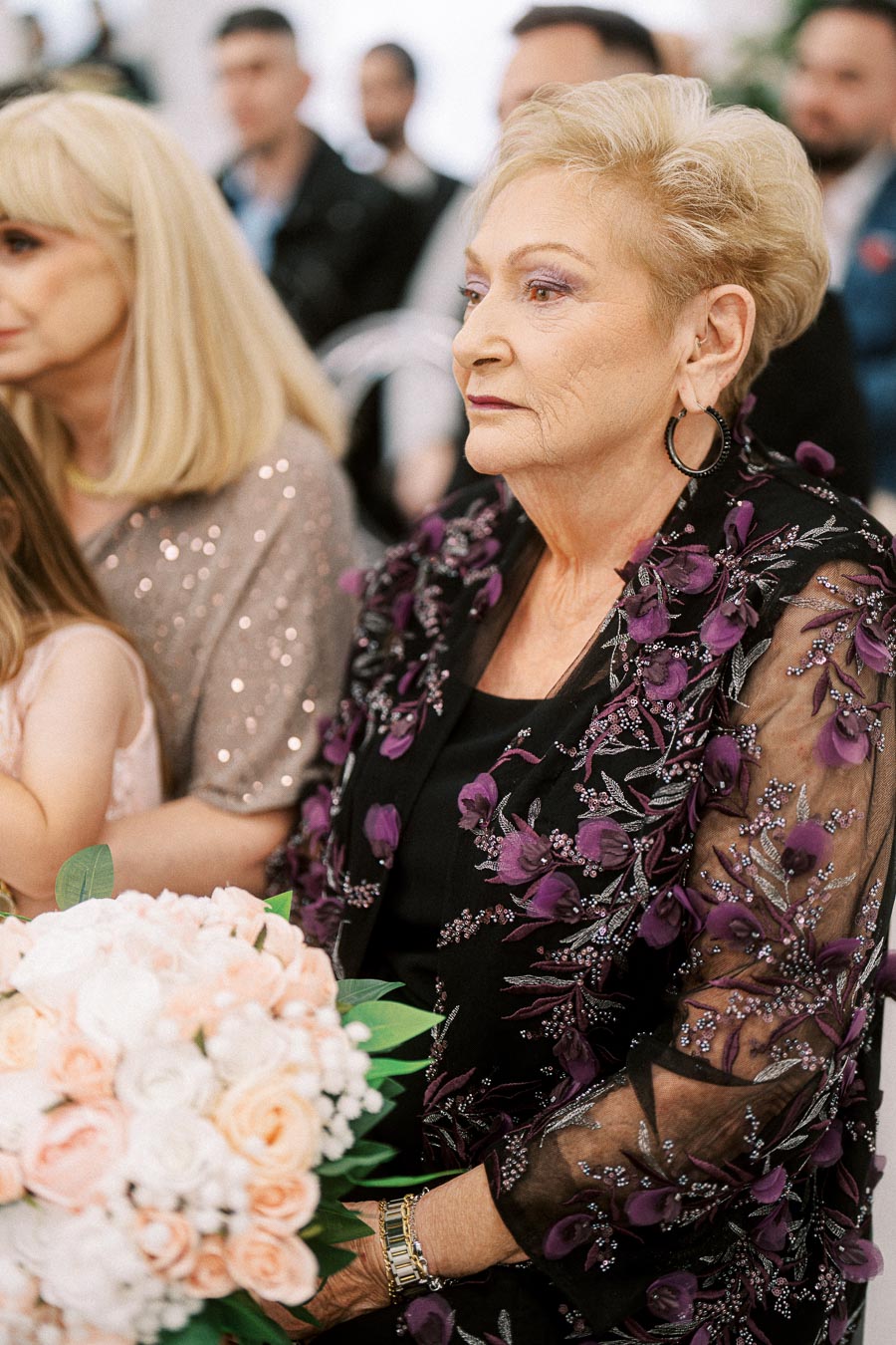 Elderly woman at formal event wearing a floral embroidered jacket, holding a bouquet of white and peach flowers, surrounded