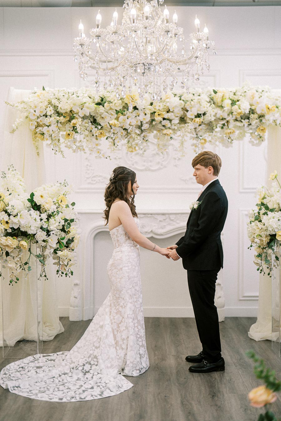 Bride and groom exchange vows under elegant floral arch and chandelier at an indoor wedding ceremony.