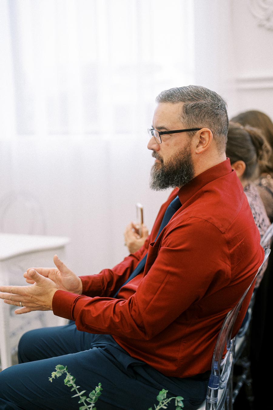 Man in a red shirt and glasses attentively listening while seated indoors at an event.