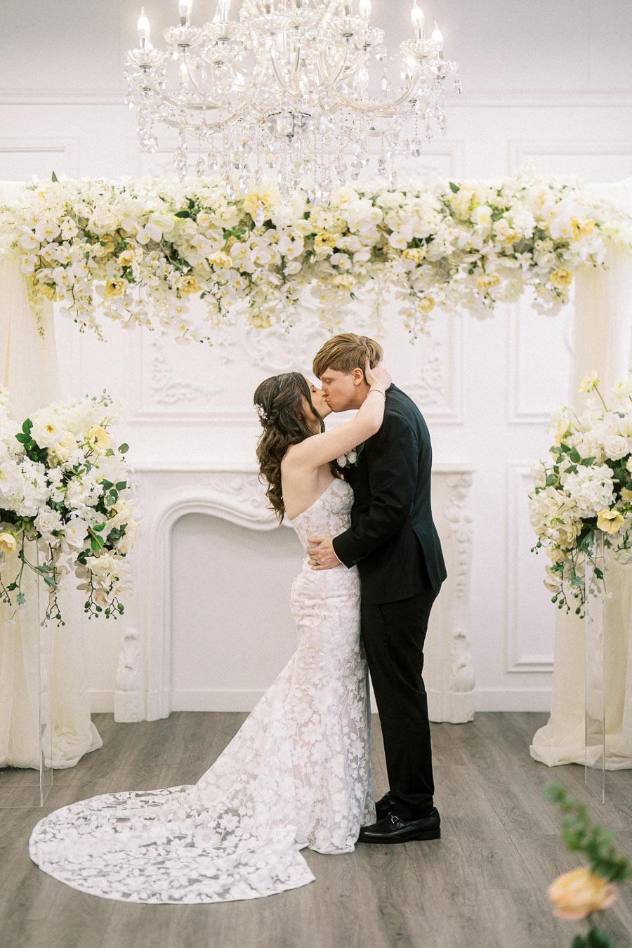 A bride and groom share a romantic kiss under an elegant chandelier, surrounded by abundant floral arrangements featuring