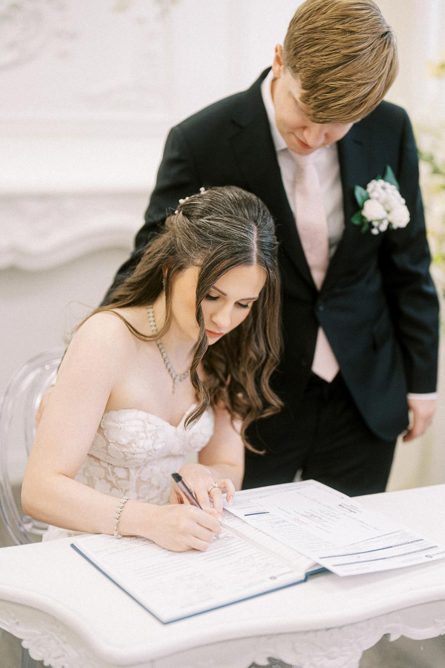 Bride signing wedding certificate beside groom in elegant ceremony setting.
