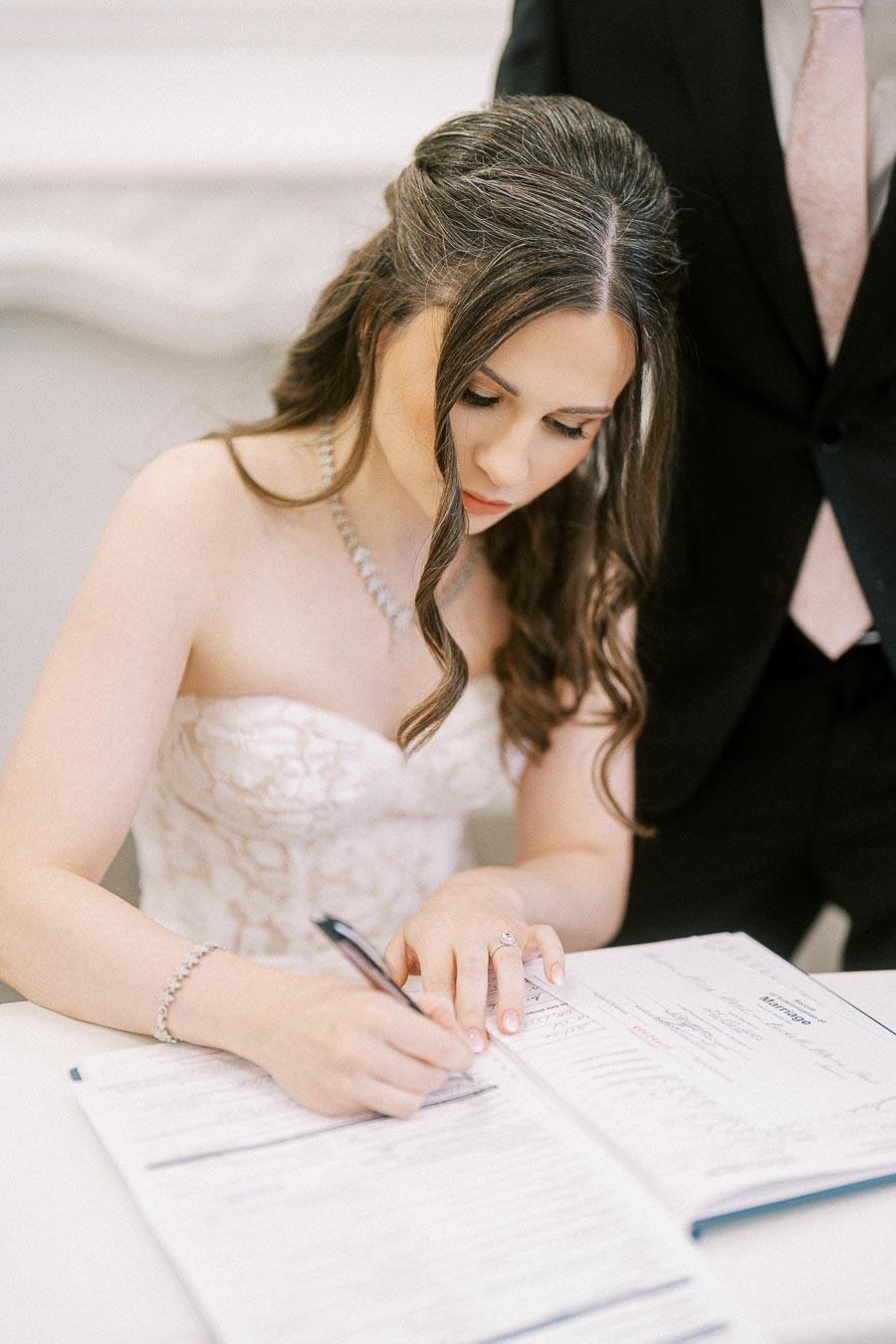 A bride signing a wedding document while wearing an elegant white lace strapless gown and sitting beside a man in a black