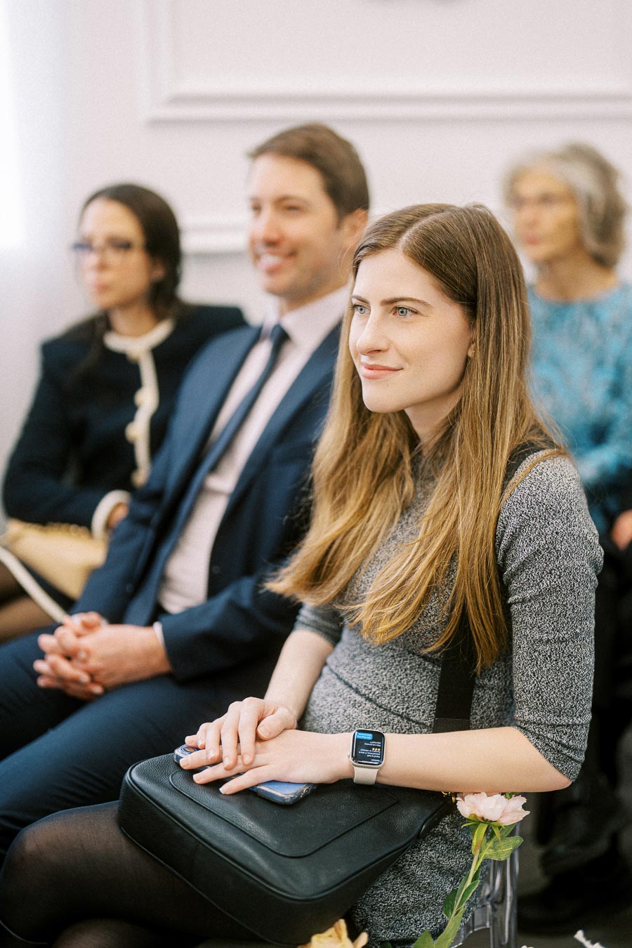 A group of attentive people sitting in a seminar, with a young woman in focus wearing a gray dress and smartwatch, holding a
