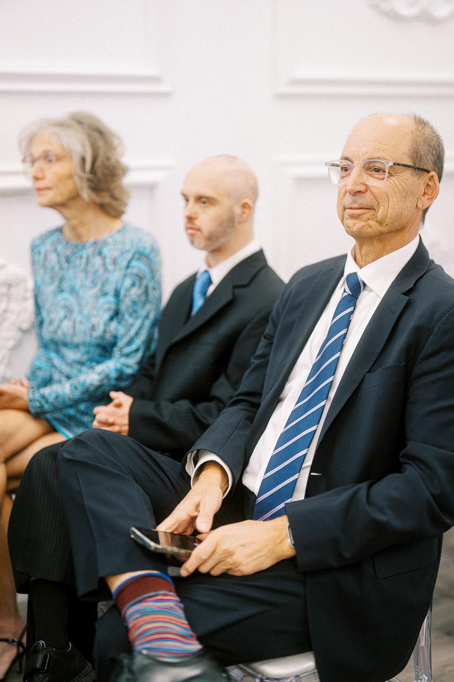 A group of people in formal attire seated at an event, with focus on a person wearing a navy suit and striped tie holding a