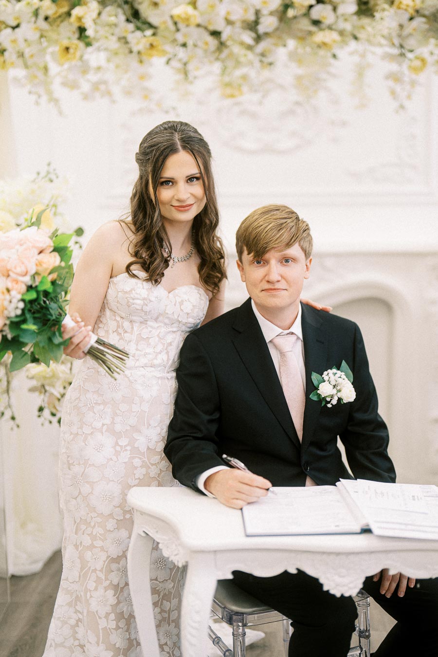 Elegant bride and groom posing at wedding ceremony, bride holding bouquet, groom signing marriage document, floral