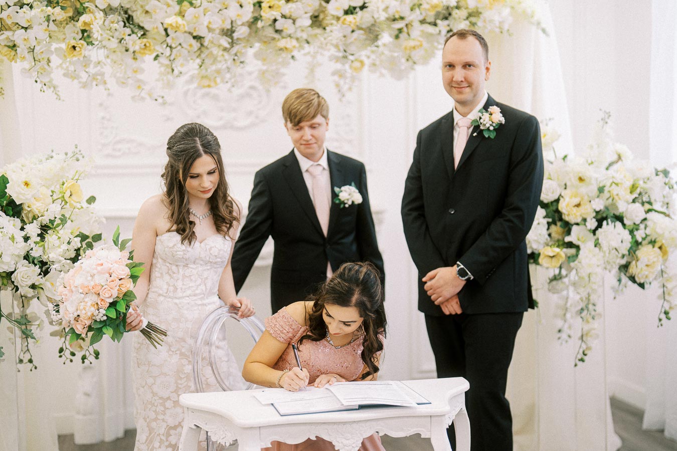 A bride in a white wedding dress and three attendants witnessing a woman in a pink dress signing a document at a wedding