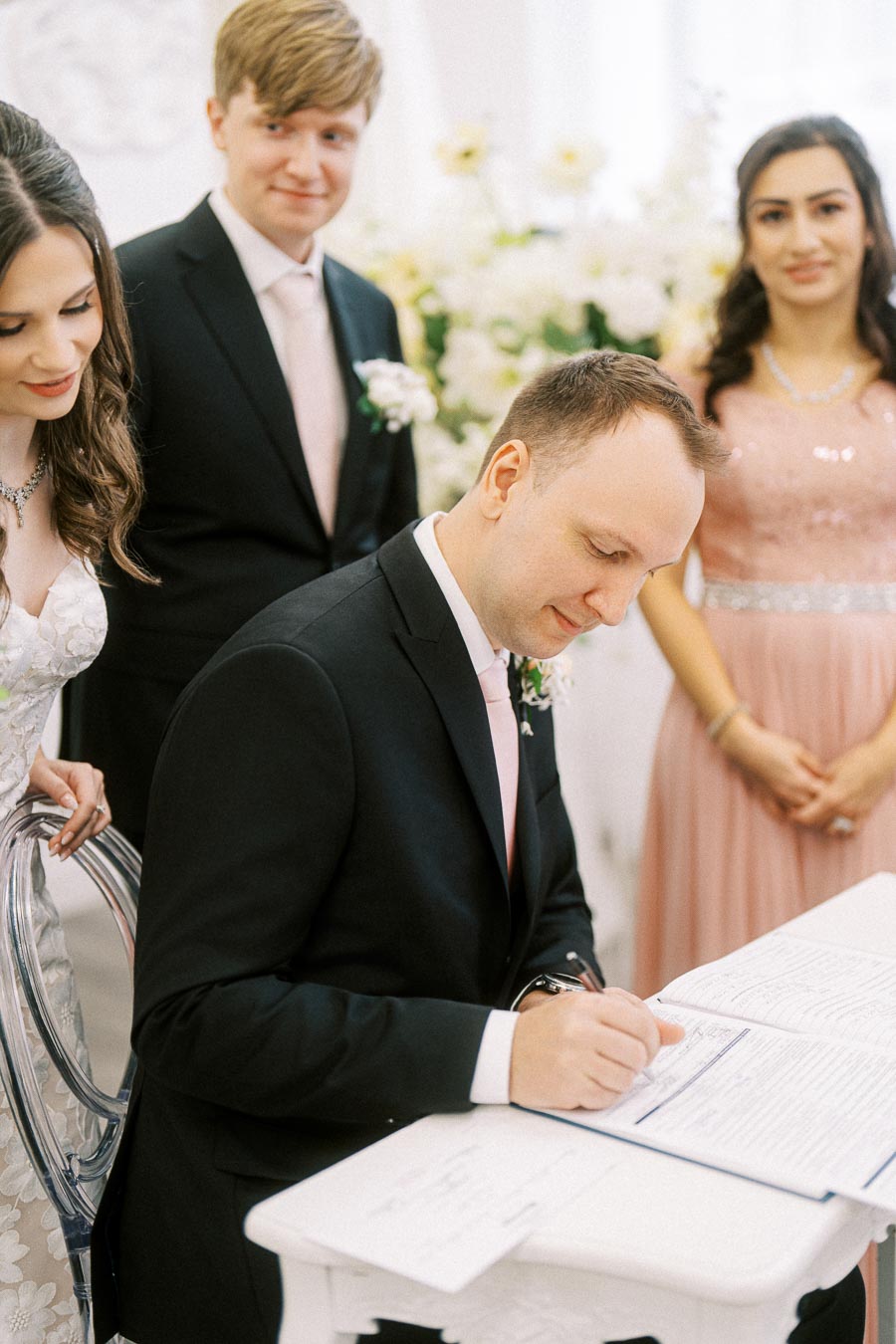 A groom signs a marriage certificate at a wedding ceremony, accompanied by the bride and two witnesses, all dressed in