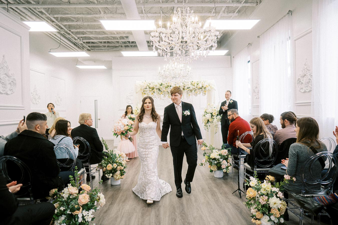 Bride and groom walking down the aisle in a beautifully decorated indoor wedding venue, surrounded by guests and floral