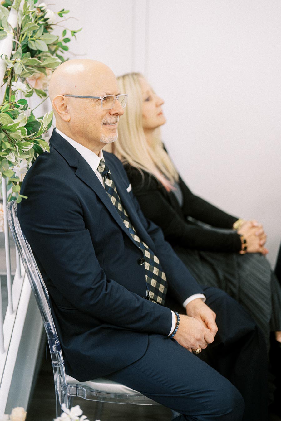 A man in a navy suit and patterned tie sits next to a woman with long blonde hair at a formal event, with decorative