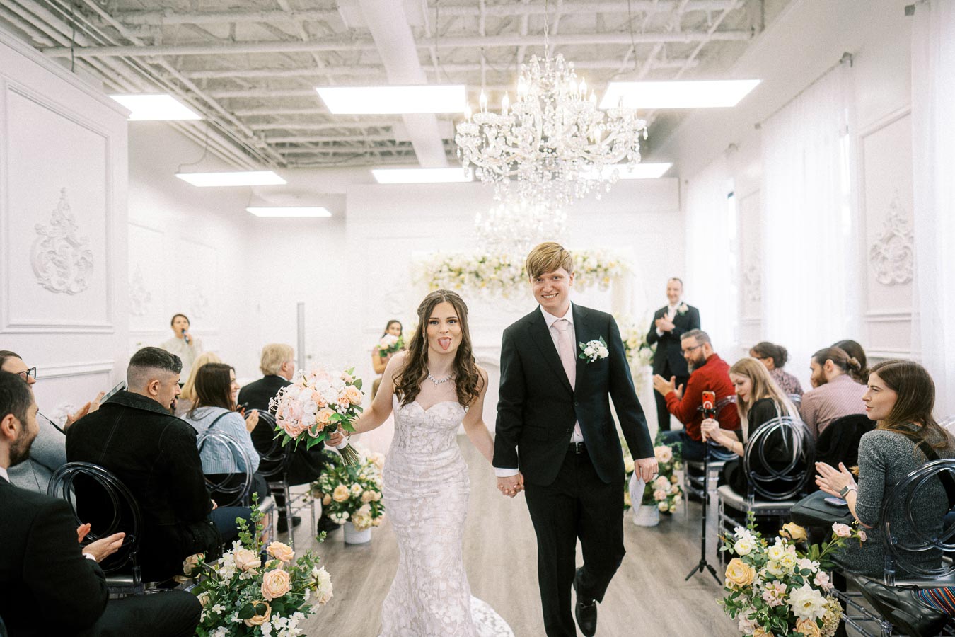 Bride and groom joyfully walking down the aisle at an elegant indoor wedding venue, with guests clapping in celebration and