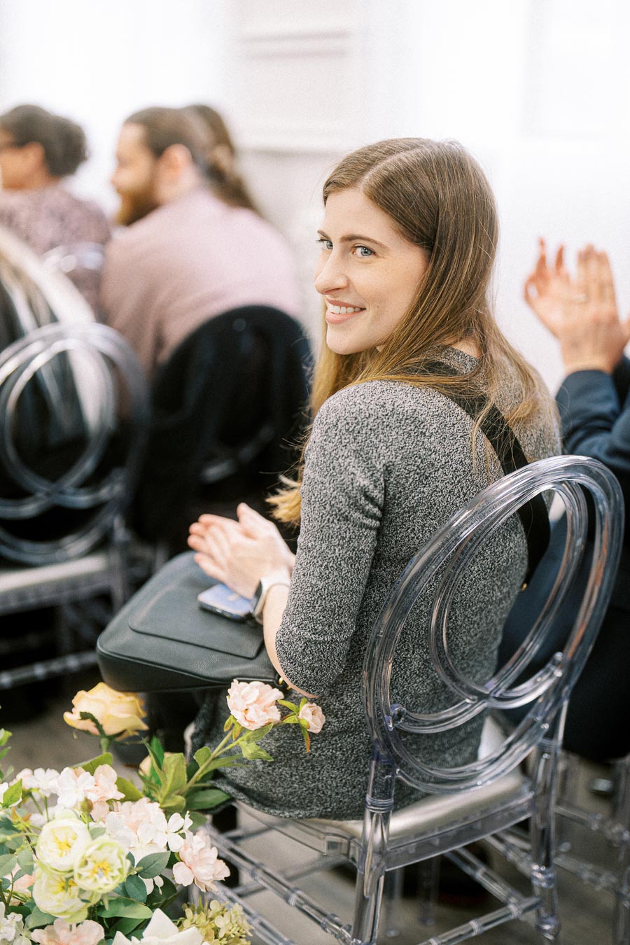 A woman in a gray sweater smiling while seated on a clear chair at an indoor event, with floral decor in the foreground and