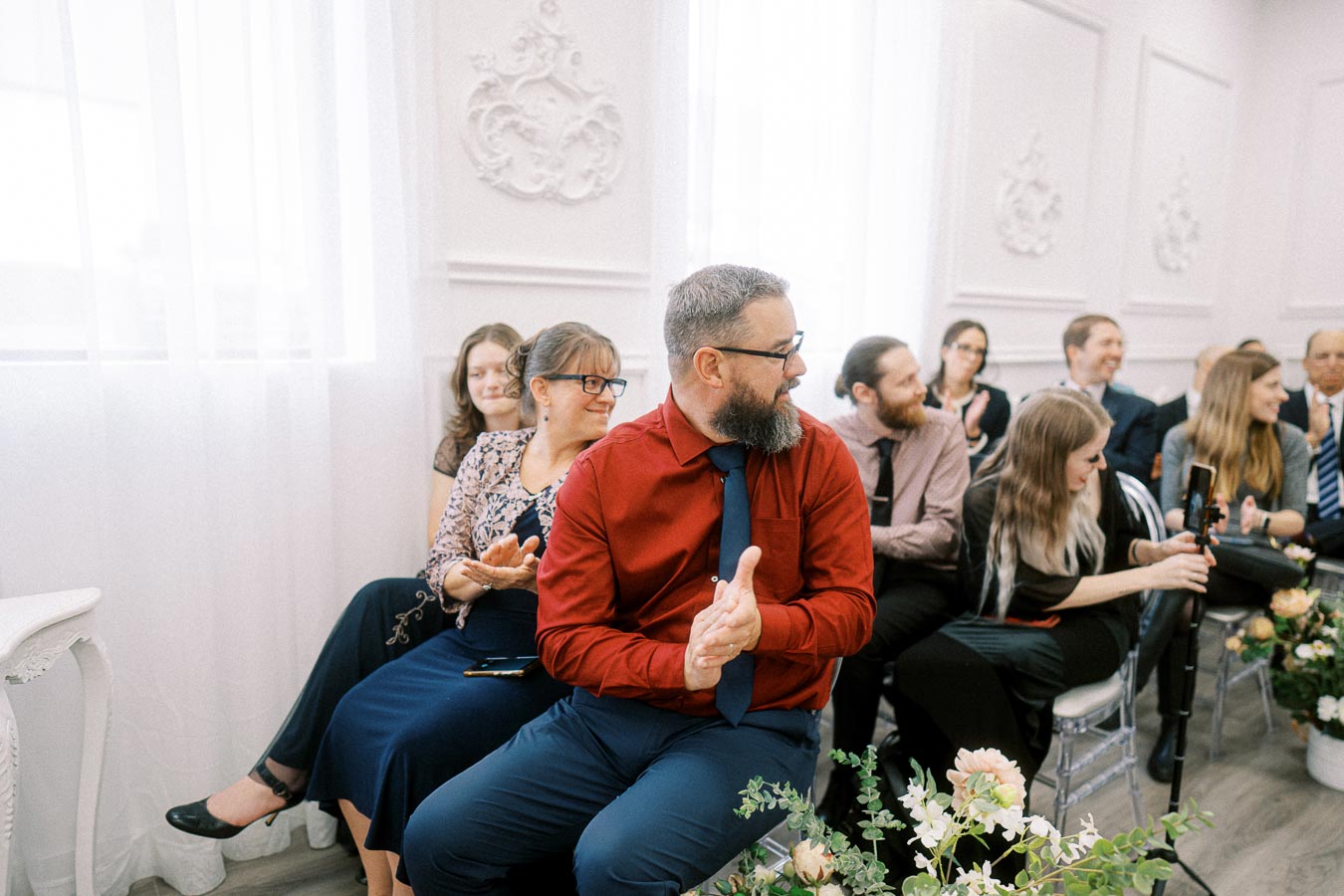 A group of people in formal attire clapping and smiling while seated in a beautifully decorated room with elegant white