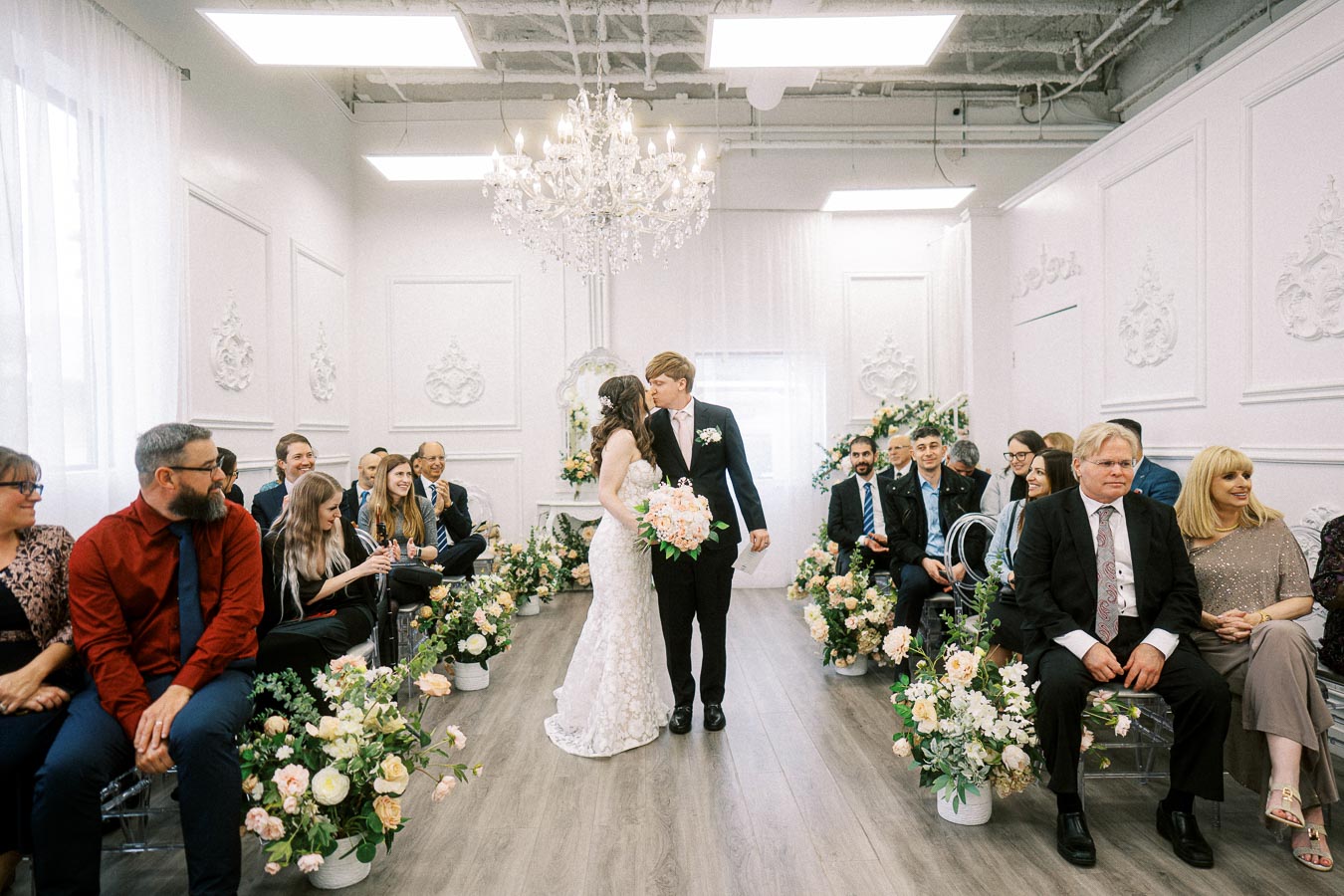 Bride and groom sharing a kiss in a beautifully decorated wedding venue, surrounded by seated guests and floral