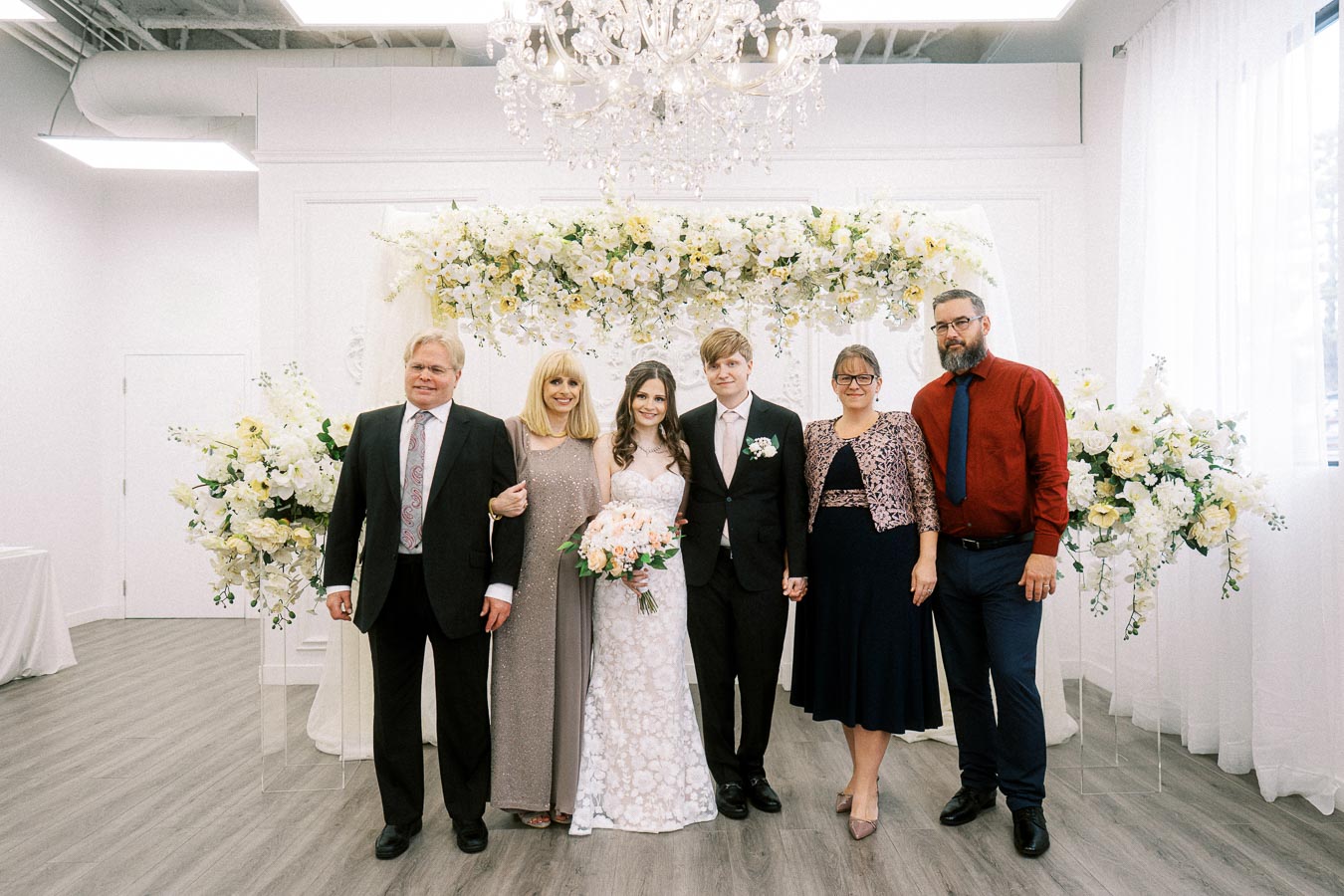 A wedding party of six poses under a floral arch. The bride, in a white lace dress, holds a bouquet of pink roses. Next to
