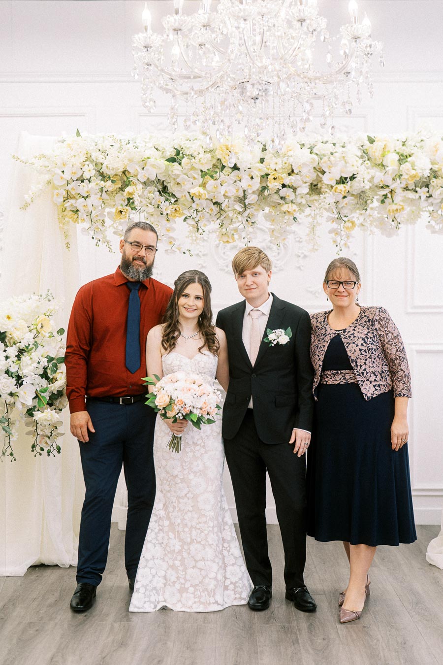 A happy wedding couple stands with two guests under a floral arch, surrounded by elegant white decor and a chandelier,