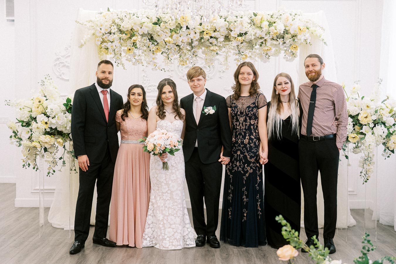 A group of seven people posing in formal attire under a floral wedding arch, with women in elegant dresses and men in suits,