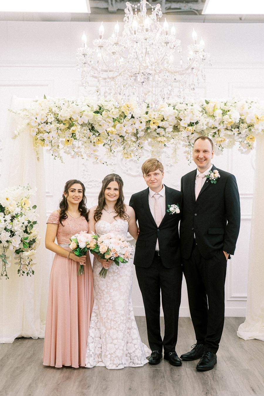 A bride and groom stand with their wedding party under a floral arch. The bride wears a white lace gown and holds a bouquet