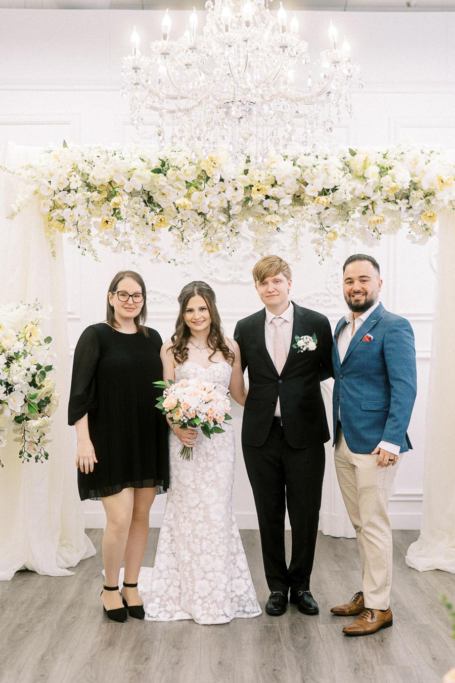Wedding group photo featuring a bride in a floral lace gown holding a bouquet, standing beside the groom in a black suit,