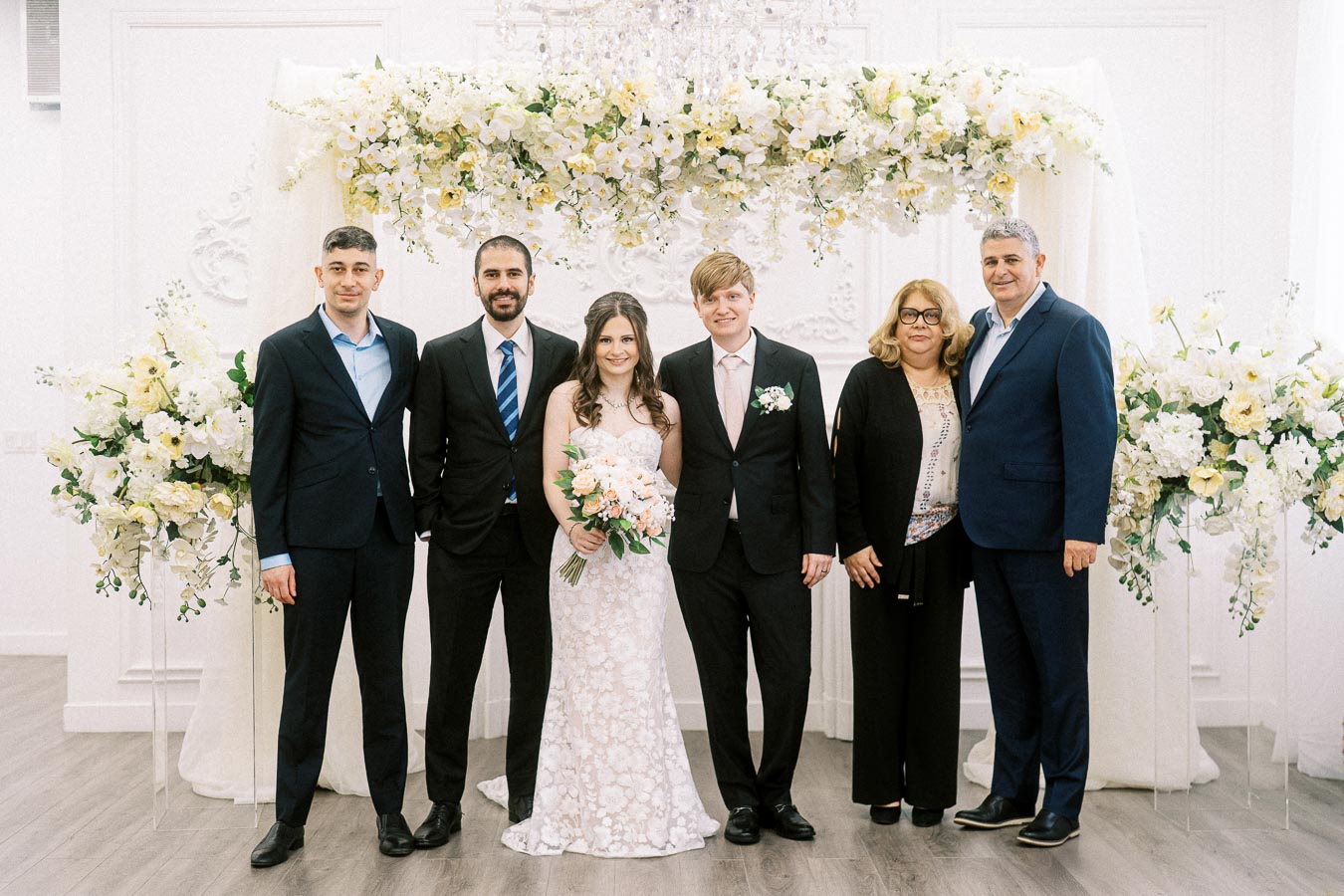 A bride and groom standing with family members in a formal wedding setting, surrounded by elegant white and yellow floral