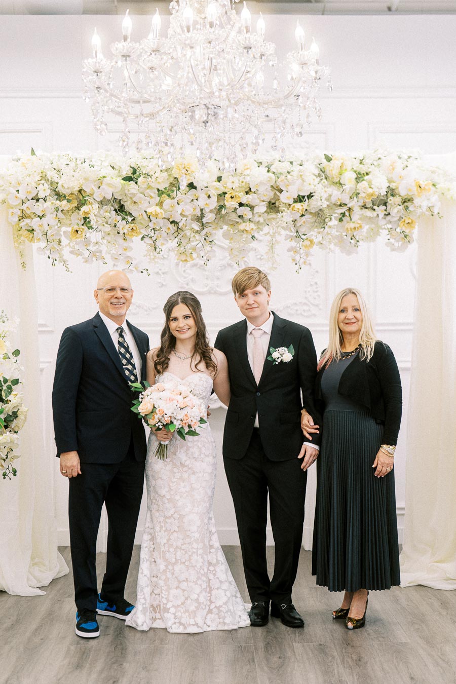 A bride and groom standing between two guests under a floral arch, with a chandelier above, at a wedding ceremony. The bride