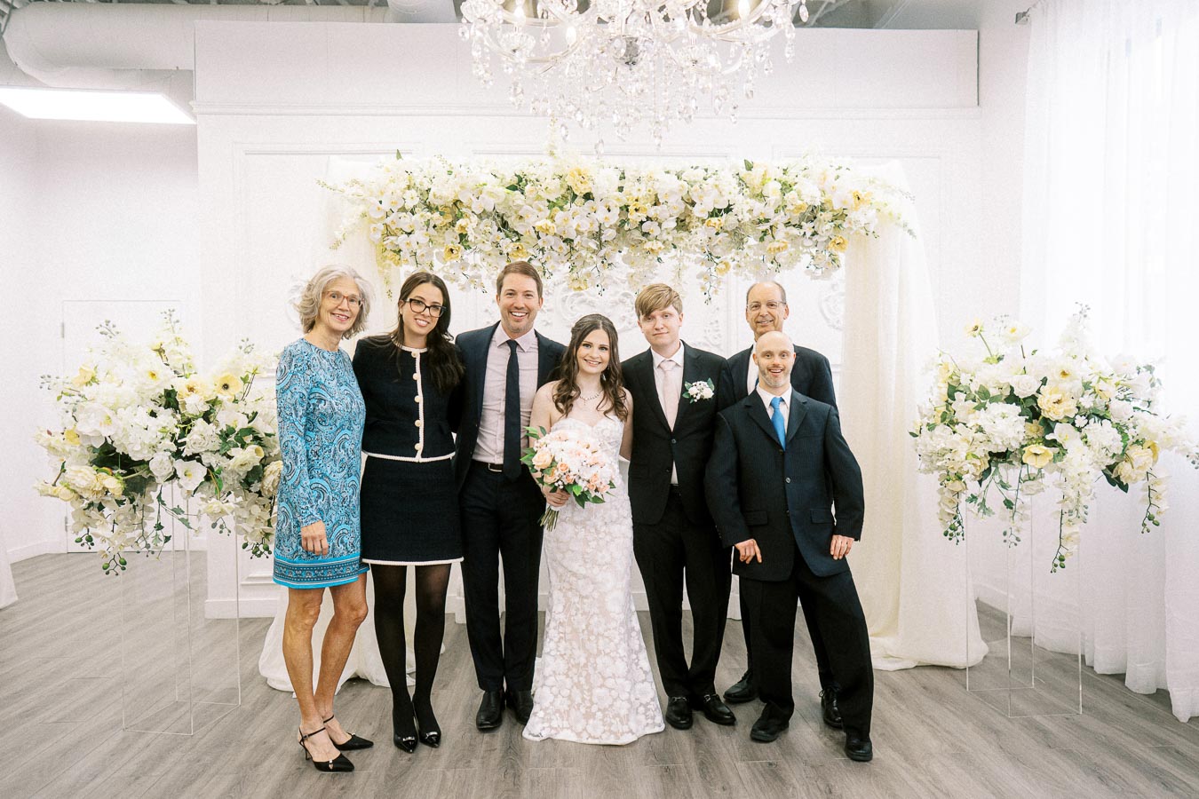 A group of people posing at a wedding ceremony, with the bride in a white dress holding a bouquet of pink flowers. The groom