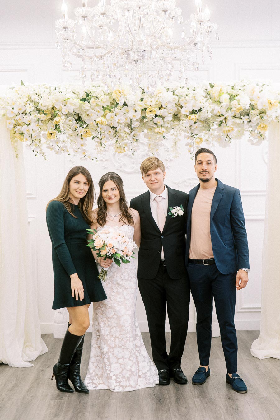 A wedding photo of a bride in a white lace dress holding a bouquet, standing alongside a groom in a black suit and two