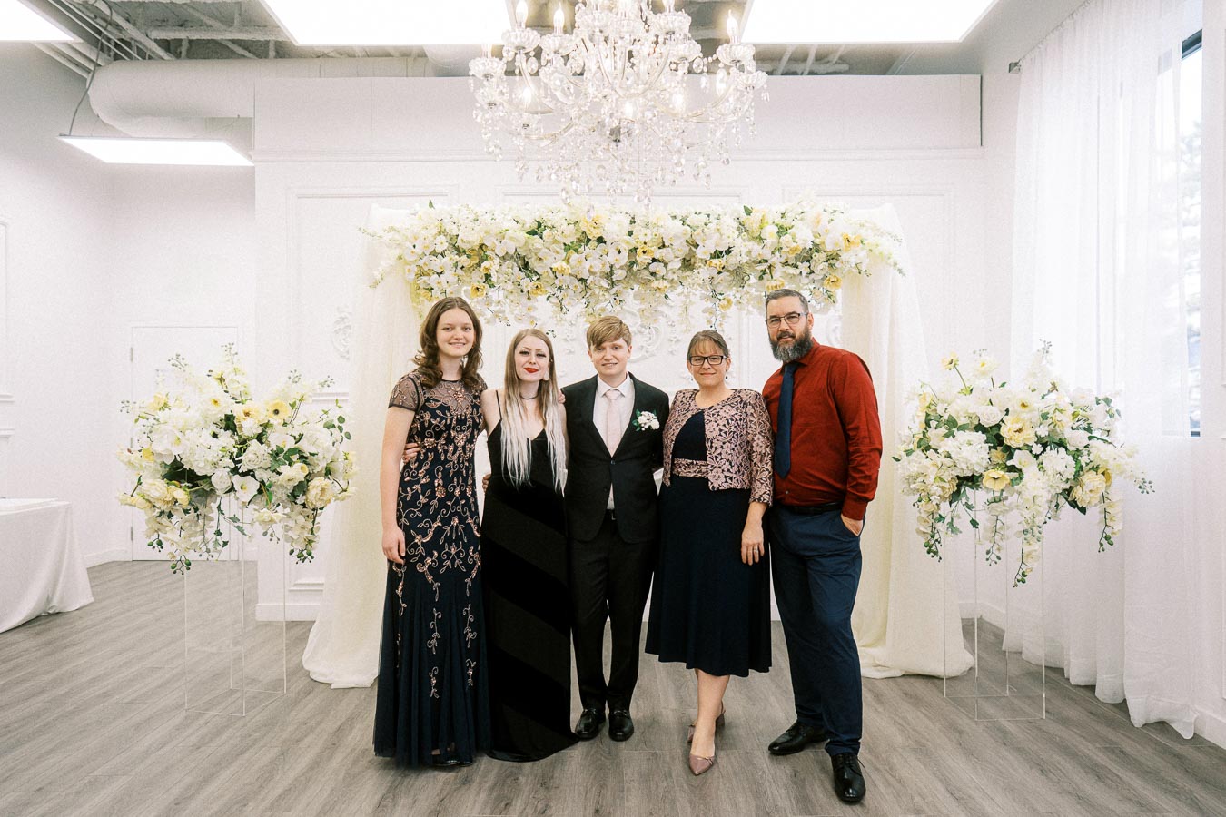 A group of five people posing at a wedding ceremony, surrounded by elegant floral arrangements and chandeliers, dressed in