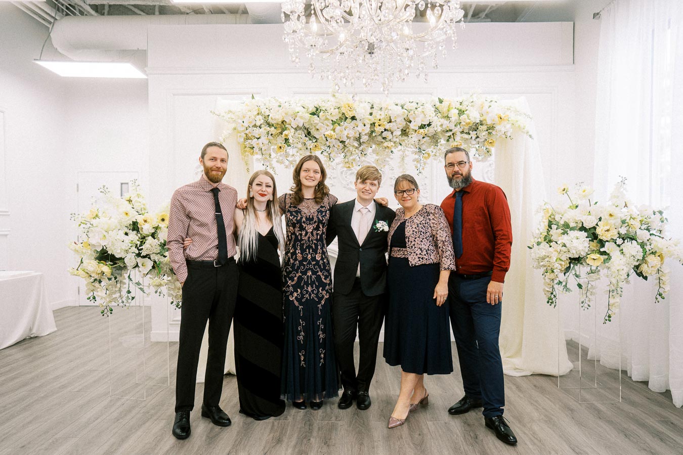 A group of people smiling in formal attire at a wedding ceremony. They are standing in front of an elegant floral arch with