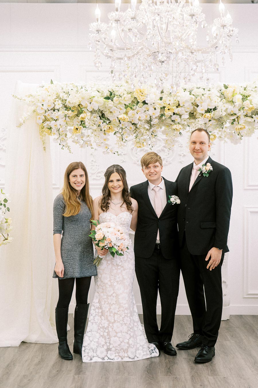 Elegant wedding portrait of a bride in a lace gown holding a pastel bouquet, flanked by guests in formal attire under a
