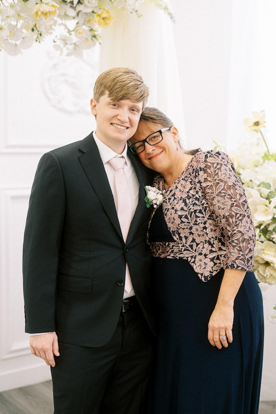 Smiling young man in a black suit with a pink tie, standing next to a woman in a navy and lace dress, posing at an elegant