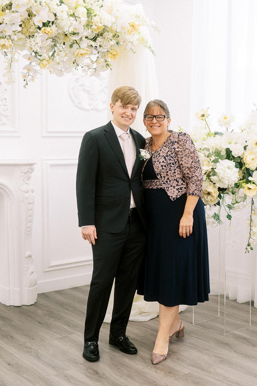 Mother and son smiling at a formal event, surrounded by elegant floral arrangements and decorative white walls.