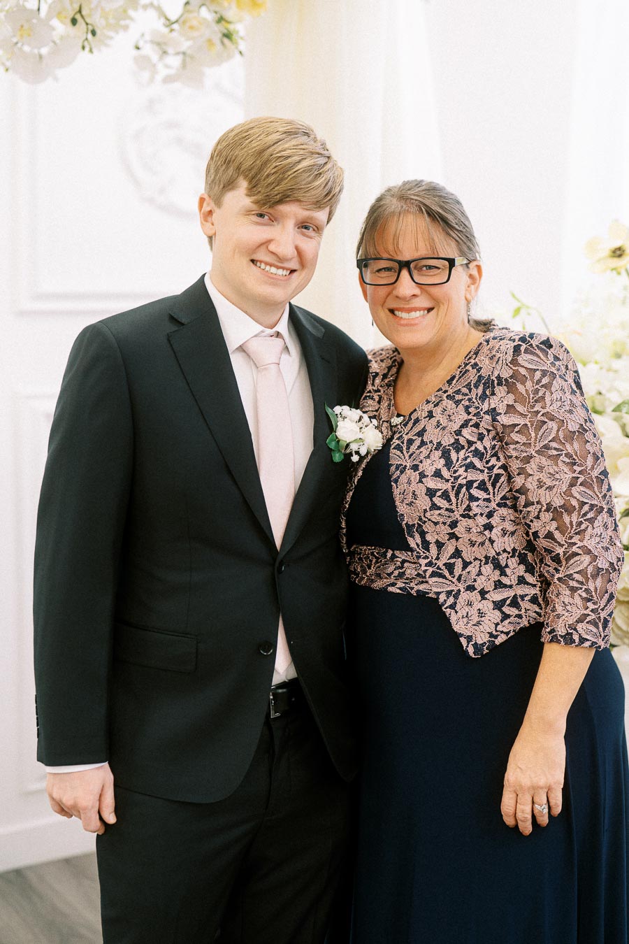 A young man in a formal black suit and pale pink tie stands smiling next to a woman wearing glasses and a navy blue dress