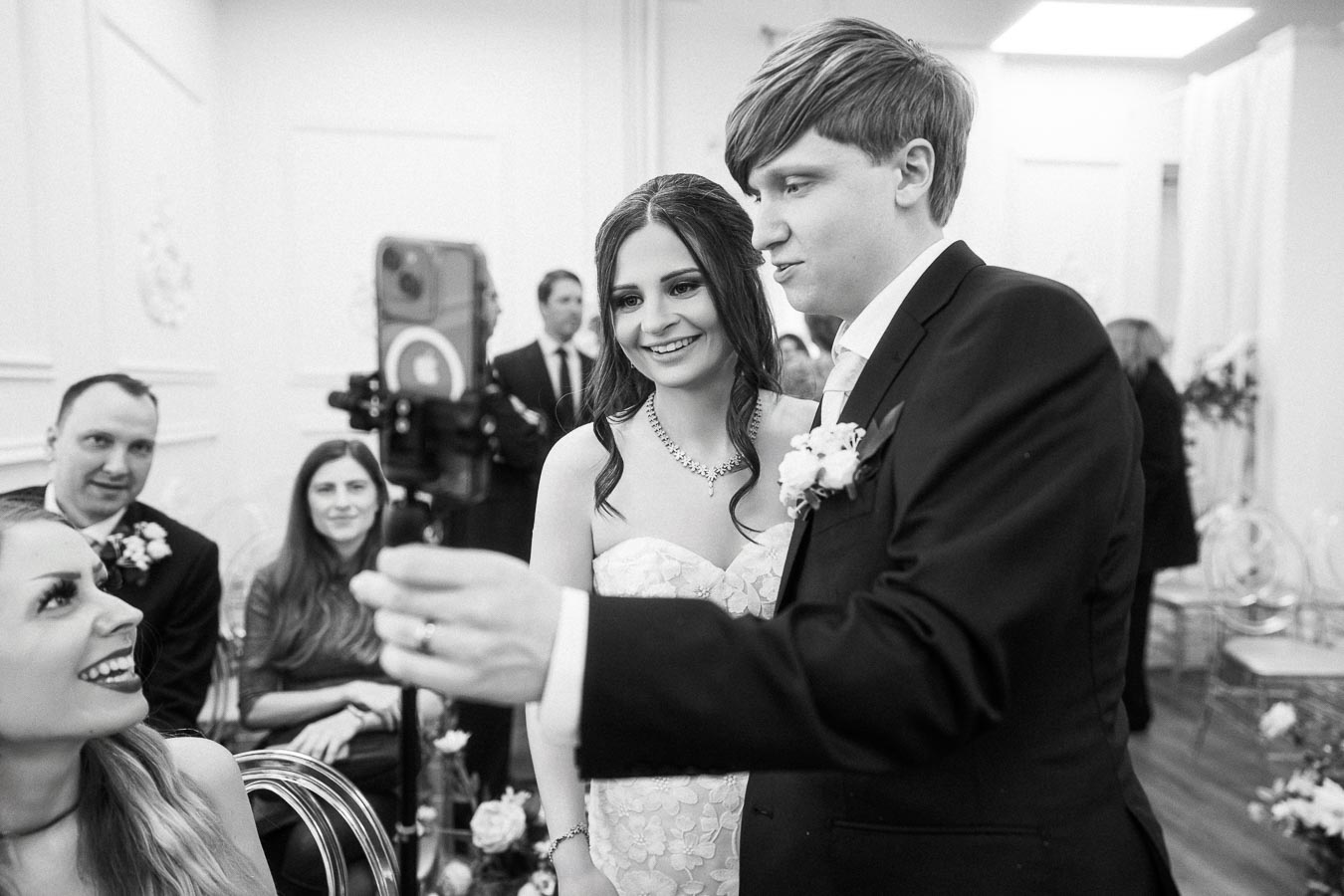 Black and white photo of a newlywed couple taking a selfie with a smartphone during their wedding ceremony, surrounded by