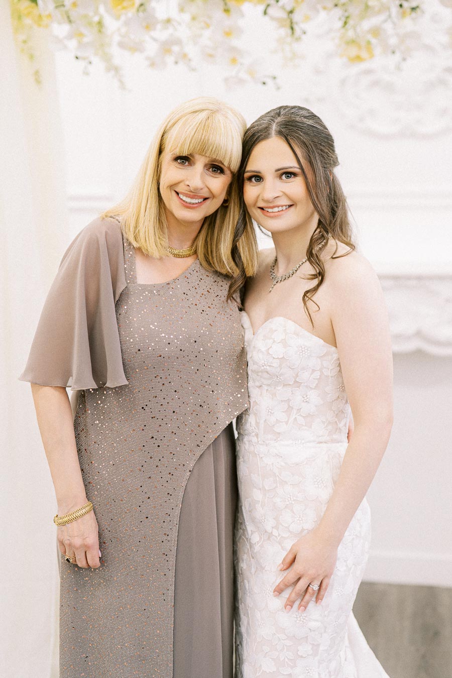 Elegant bridal portrait featuring bride in lace gown standing with smiling woman in sparkling formal dress, set against a