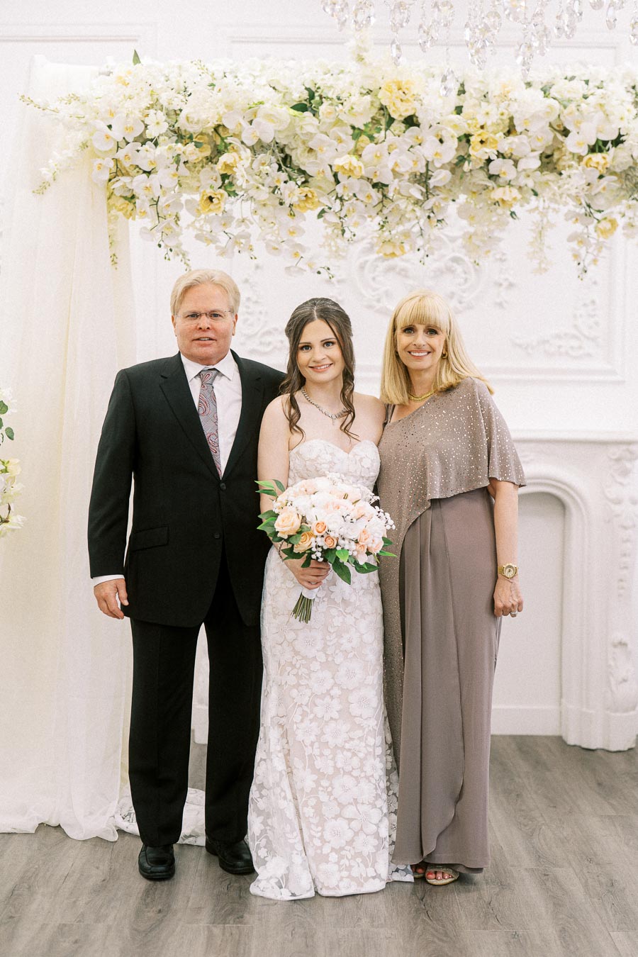 A bride in a white lace wedding dress stands between two elegantly dressed guests, under a floral arch with white and yellow